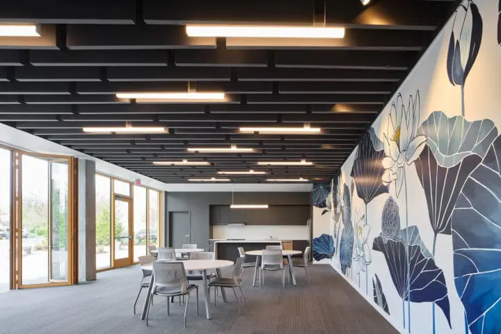 View of 72Foster’s ground-floor community room, which features a kitchen, several tables, gray acoustic panels hanging from the ceiling, and a vibrant blue floral mural.