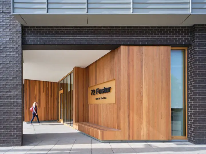 A person carrying a shoulder bag walks toward 72Foster’s front door, past a built-in wooden bench, which is situated under the “72Foster” sign.