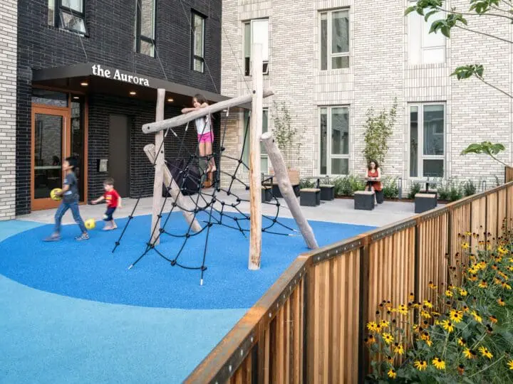 Three kids play in a fenced-in outdoor play space at The Aurora while a person sits at one of the outdoor tables to supervise them.
