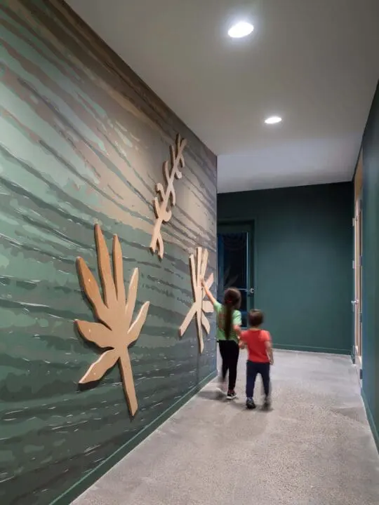 Two kids walk down a deep teal hallway holding hands and touching one of the three leaf motifs on the wall at The Aurora.