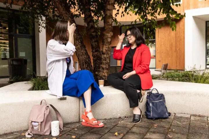 Two people sit facing each other on a curved bench, communicating in ASL.
